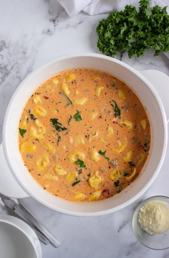 A white pot filled with creamy tomato tortellini soup with greens, placed on a marble surface next to kale and a small dish of grated cheese.