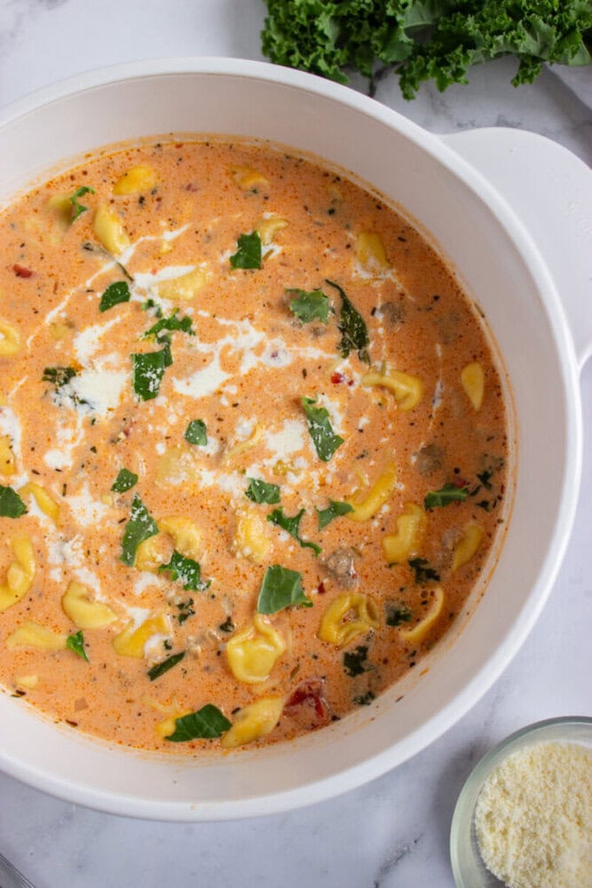 A pot of creamy tortellini soup with herbs and greens, seen from above on a marble countertop.