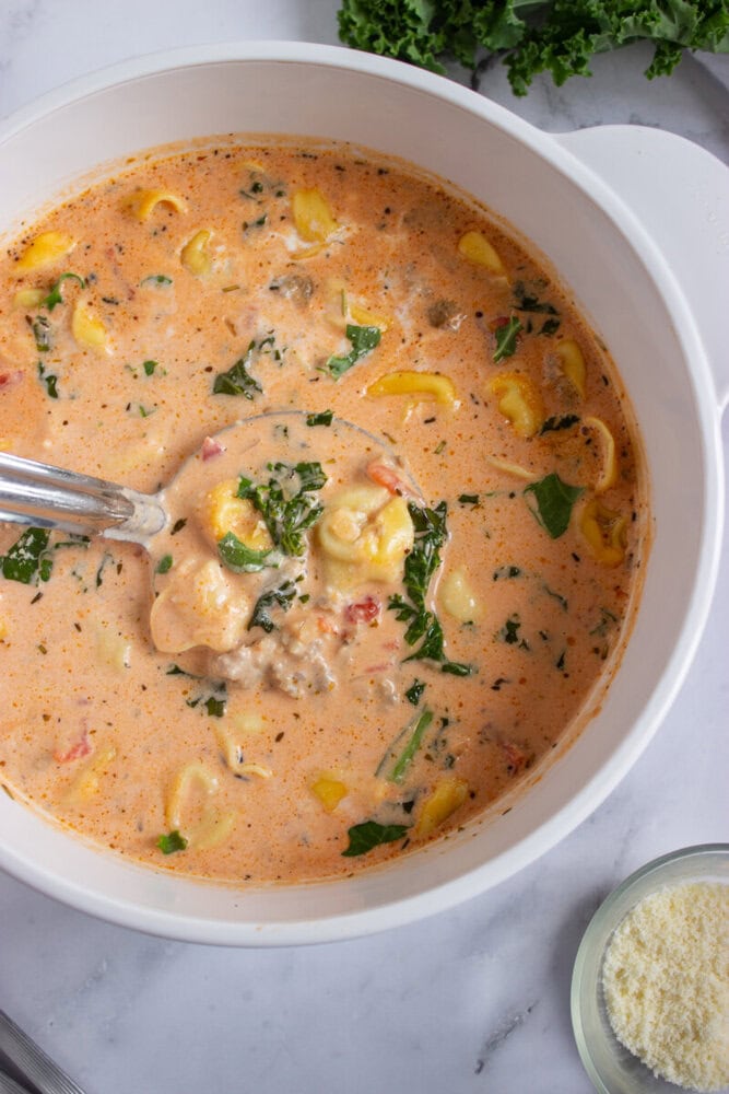 A white pot filled with creamy soup containing tortellini, sausage, chopped greens, and herbs, with a metal ladle inside. A bowl of grated cheese sits nearby.