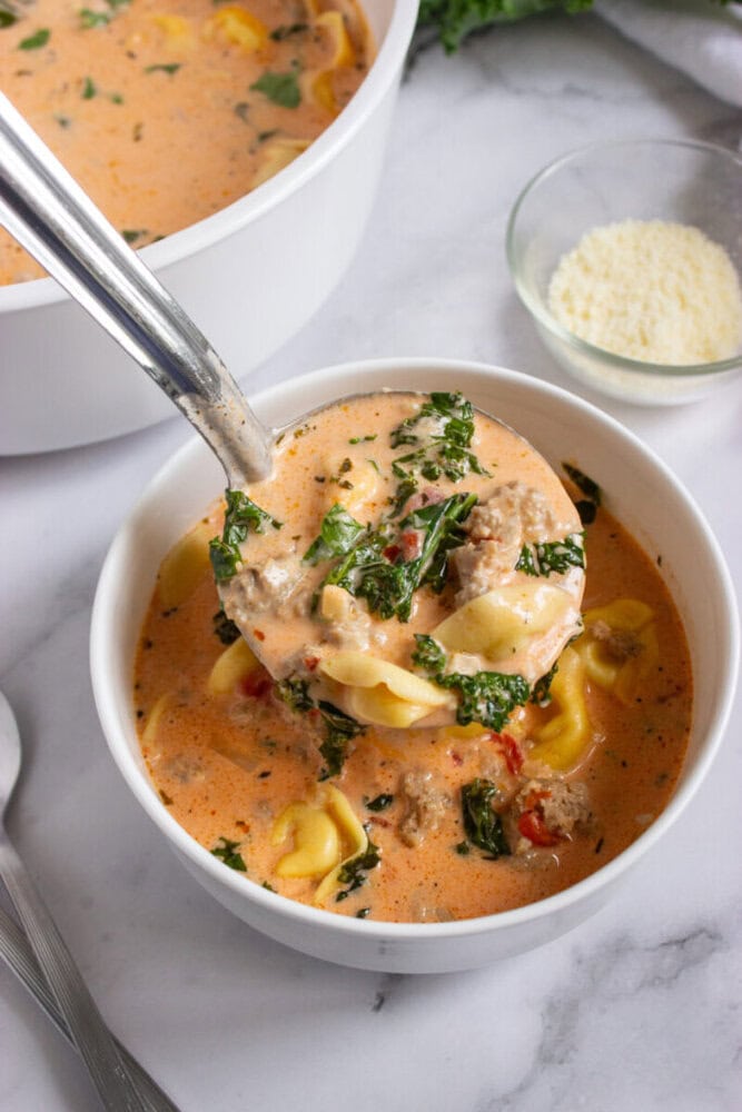 A ladle serving creamy soup with tortellini, sausage, kale, and herbs into a white bowl, with grated cheese in a small glass dish nearby.