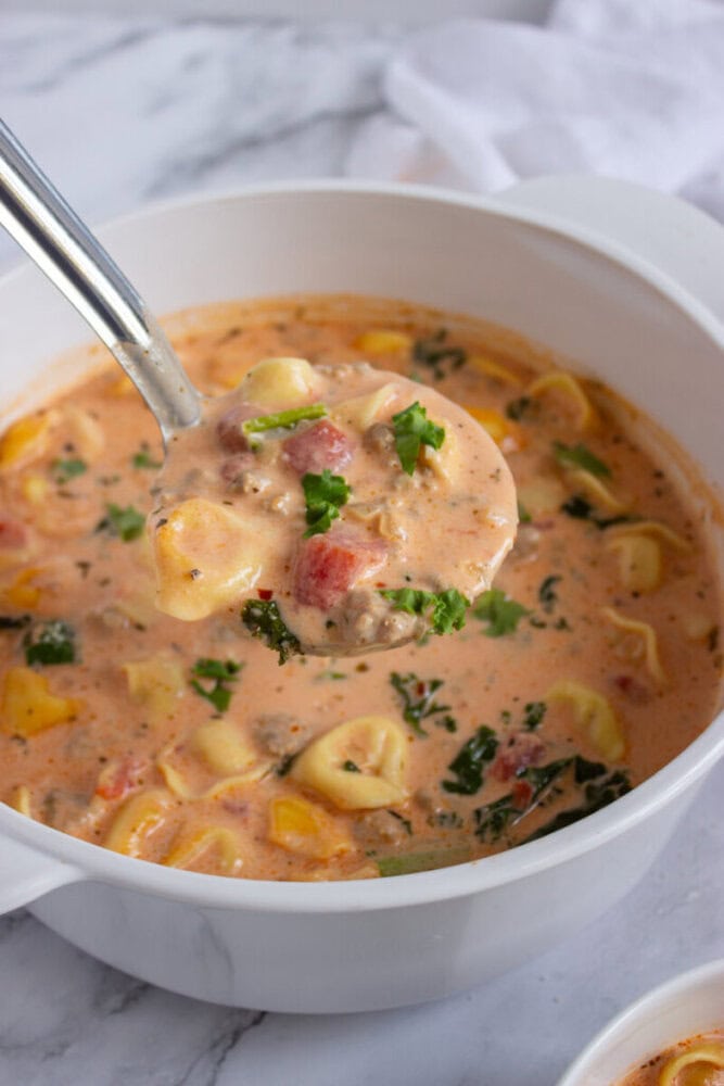 A ladle lifts creamy tortellini soup with tomatoes, spinach, and herbs from a white pot on a marble surface.
