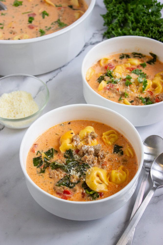 Two white bowls of creamy tortellini soup with sausage, spinach, and tomatoes on a marble surface, next to a bowl of grated cheese, fresh kale, and metal spoons.