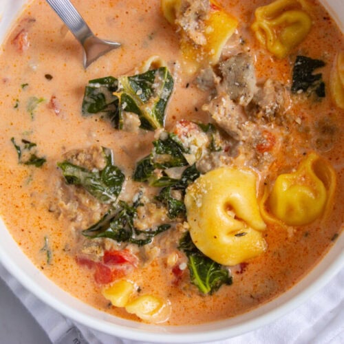 A bowl of creamy soup with tortellini, ground sausage, kale, and tomatoes, with a spoon resting inside.