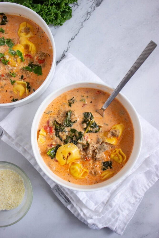A bowl of creamy soup with tortellini, ground meat, kale, and tomatoes, served with a spoon; another bowl and a small dish of grated cheese are nearby.