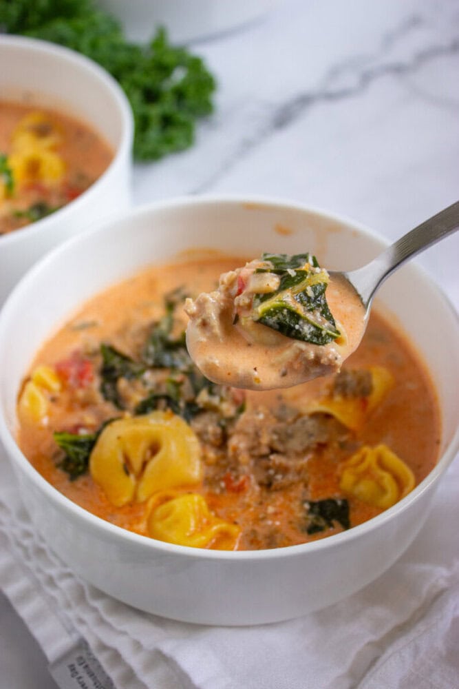 A bowl of creamy tortellini soup with sausage, kale, and tomatoes. A spoon is lifted, showing the soup’s ingredients.