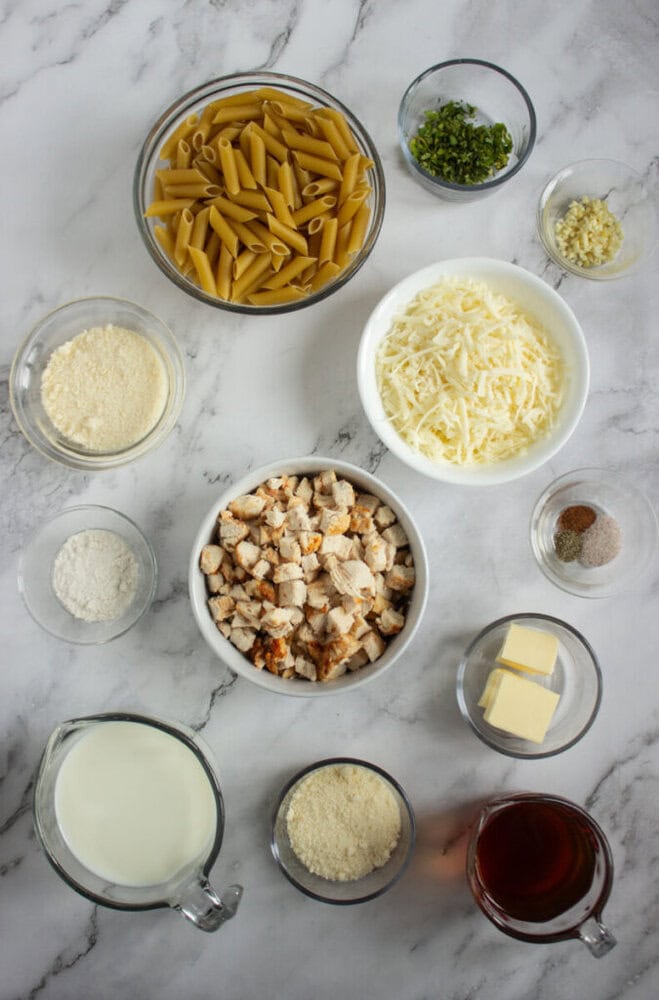 Various ingredients for pasta are arranged on a marble surface, including uncooked penne, cooked chicken, shredded cheese, milk, butter, flour, seasonings, and broth in glass bowls.