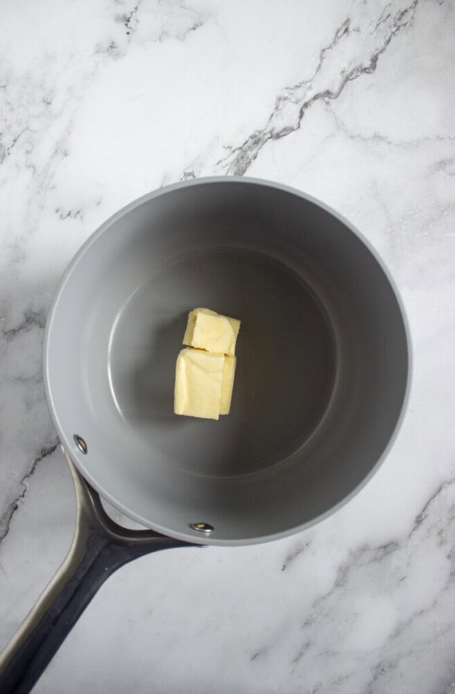 A metal saucepan with two cubes of butter inside sits on a white marble countertop.