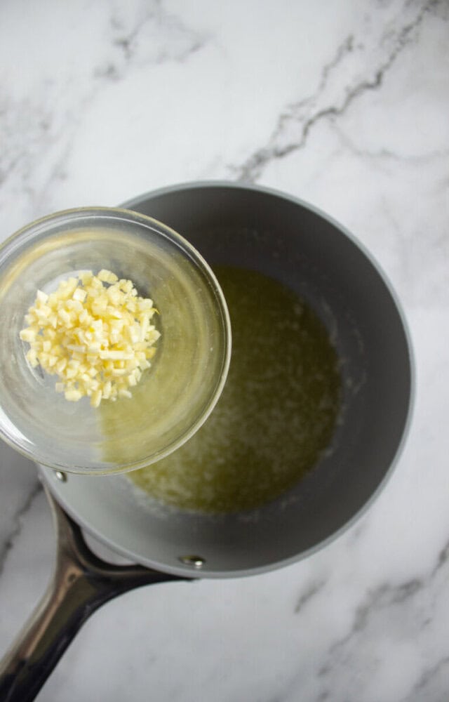 A small glass bowl of chopped garlic is held above a saucepan containing melted butter on a marble countertop.