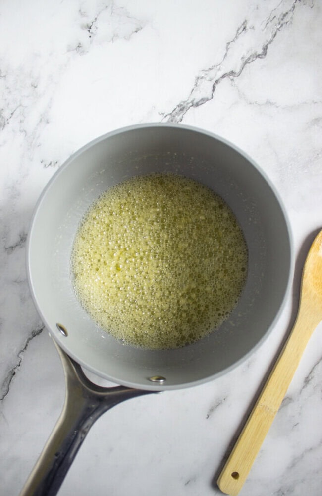 A saucepan with melted butter or a bubbling liquid sits on a marble countertop next to a wooden spoon.