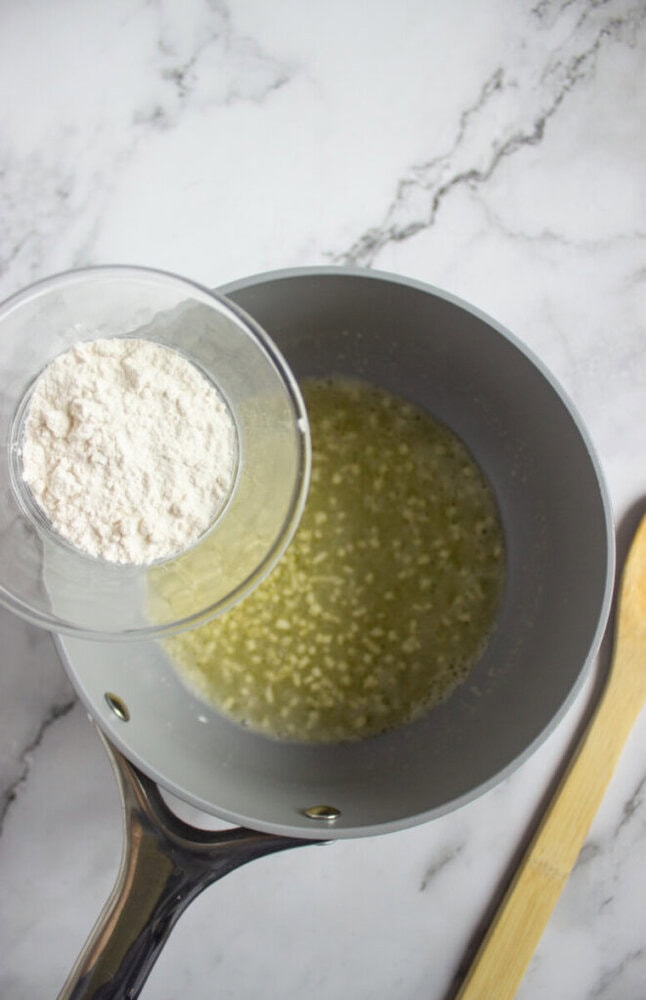 A glass bowl of flour is being added to a saucepan of melted butter on a marble countertop, with a wooden spoon beside the pan.