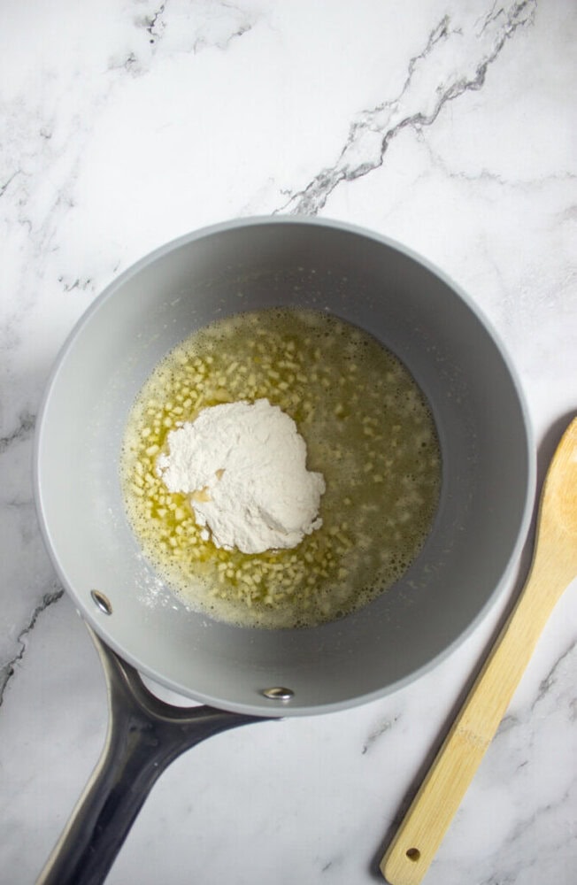 A saucepan with melted butter and a mound of flour inside, sitting on a marble countertop next to a wooden spatula.