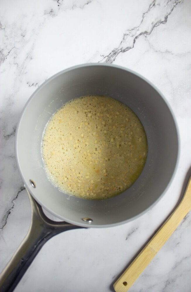 A saucepan with a bubbly, pale yellow mixture sits on a marble surface next to a wooden spoon.