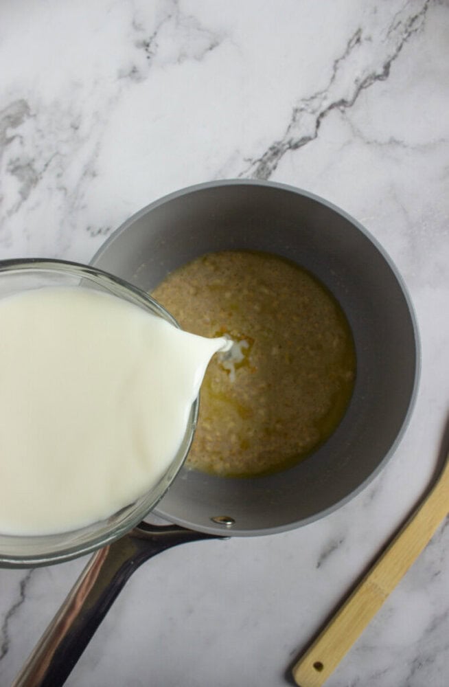 A glass measuring cup pours milk into a saucepan containing a beige mixture, on a marble countertop with a wooden spoon nearby.