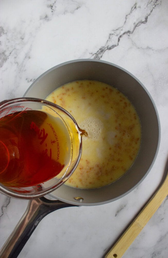 A measuring cup pours a brown liquid into a saucepan containing a yellow mixture, set on a marble countertop beside a wooden spoon.