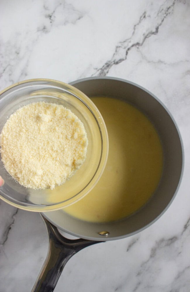 A hand holds a bowl of grated cheese above a saucepan filled with a yellow liquid mixture on a marble countertop.