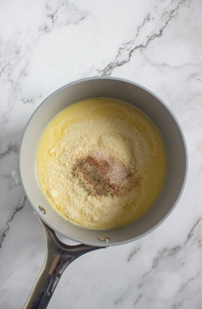 A saucepan on a marble surface containing melted butter, grated cheese, and a sprinkle of spices.