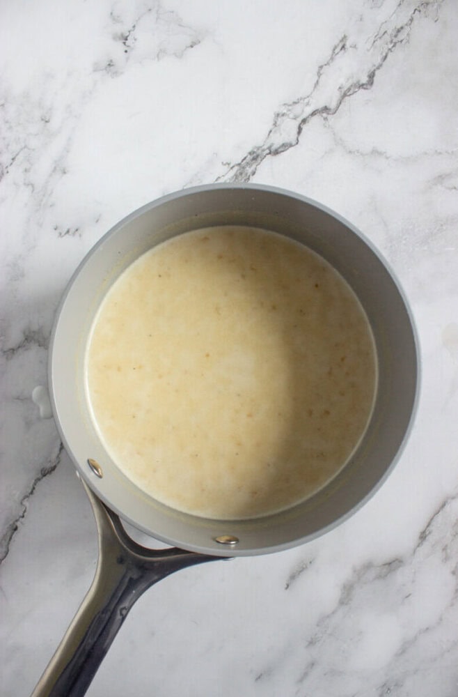 A saucepan containing a light beige liquid mixture sits on a white marble surface.