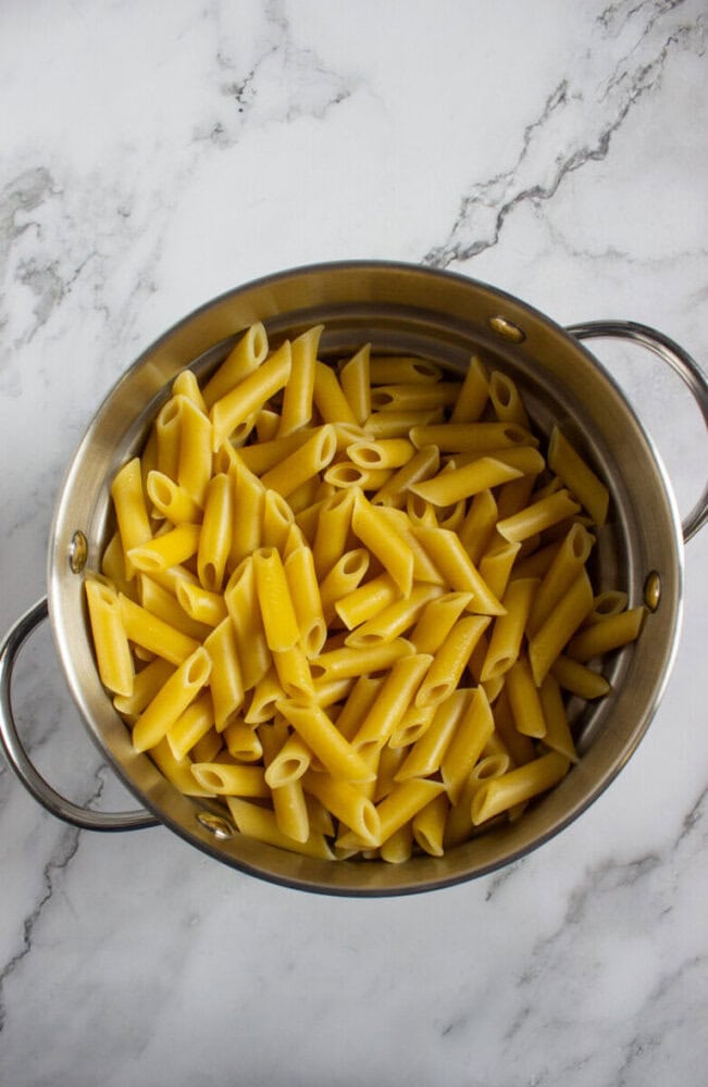 A stainless steel pot filled with cooked penne pasta sits on a white marble surface.