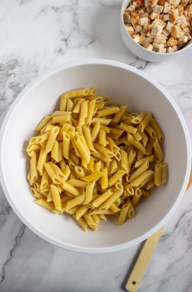 A white bowl filled with cooked penne pasta sits on a marble surface, with a spoon and a separate bowl of diced cooked chicken nearby.