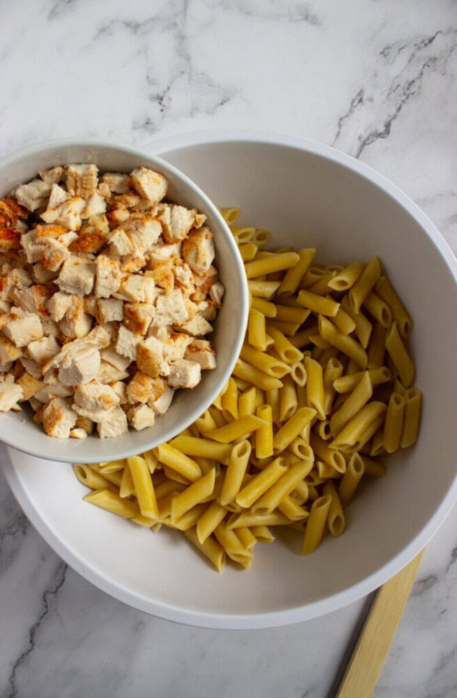 A large white bowl with cooked penne pasta and a smaller bowl of diced cooked chicken being poured into it, on a marble surface.