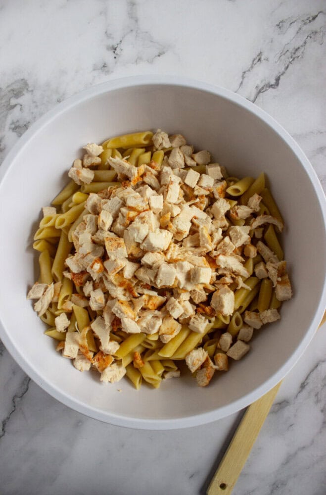 A white bowl filled with cooked penne pasta and diced cooked chicken sits on a marble countertop.