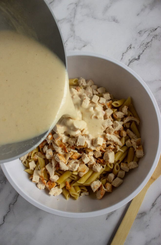 A creamy sauce is being poured from a pot over chopped cooked chicken and penne pasta in a white mixing bowl on a marble countertop.