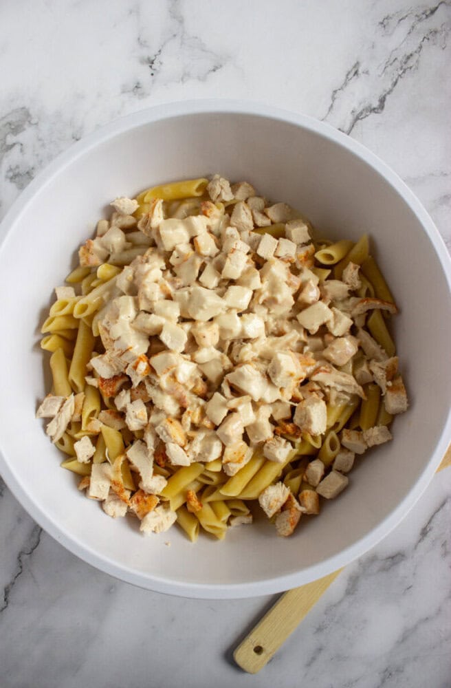 A white bowl containing cooked penne pasta topped with chunks of cooked chicken and a creamy sauce, placed on a marble surface.