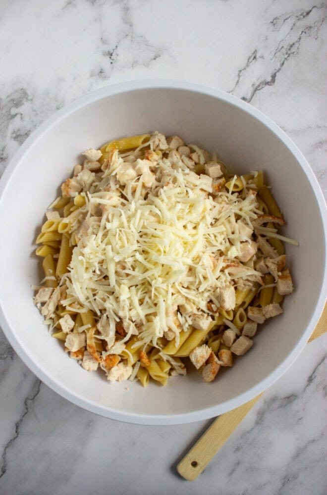 A white bowl on a marble surface contains penne pasta, diced cooked chicken, and shredded cheese. A wooden spoon rests beside the bowl.