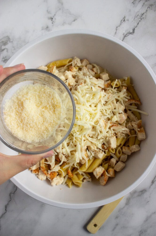 A bowl of cooked penne pasta, diced chicken, and shredded cheese, with a hand holding a small bowl of grated cheese above it.