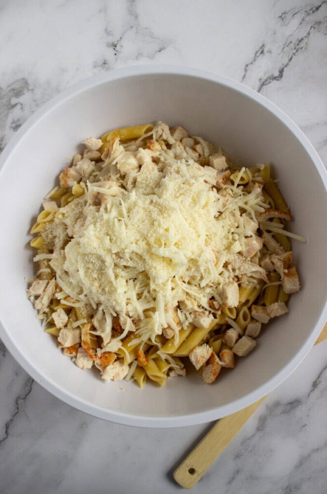 A white bowl filled with penne pasta, diced cooked chicken, and shredded cheese on a marble countertop.