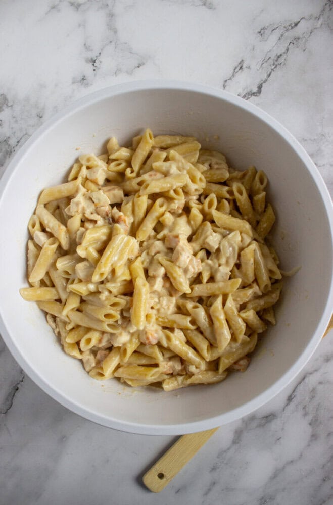 A white bowl filled with creamy penne pasta, mixed with small pieces of meat or seafood, on a marble countertop.
