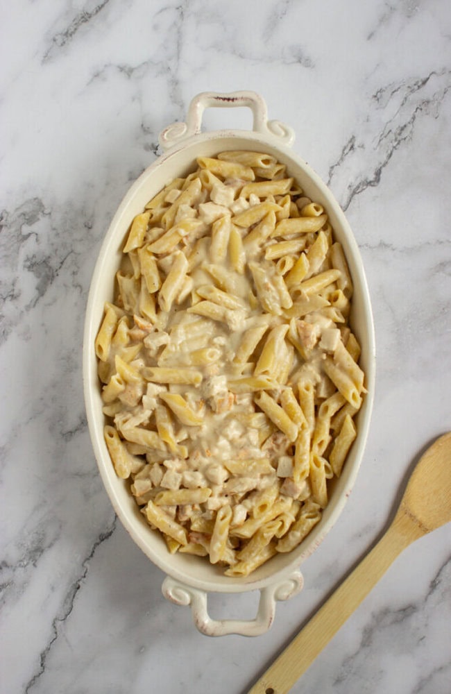 An oval white baking dish filled with penne pasta in a creamy white sauce sits on a marble surface next to a wooden spoon.