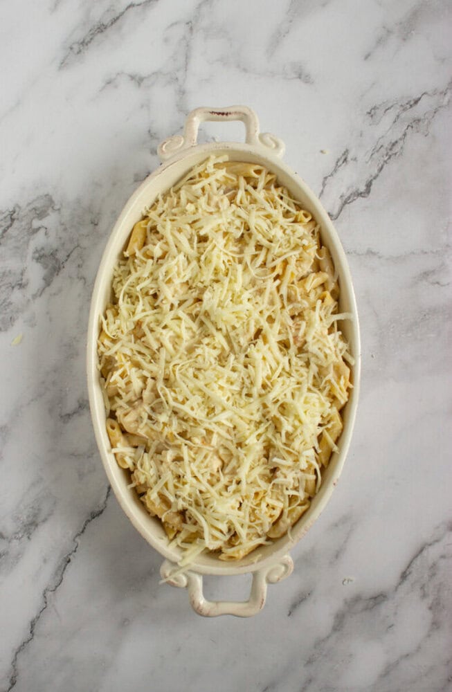 An oval baking dish filled with uncooked pasta covered with shredded cheese, placed on a marble countertop.