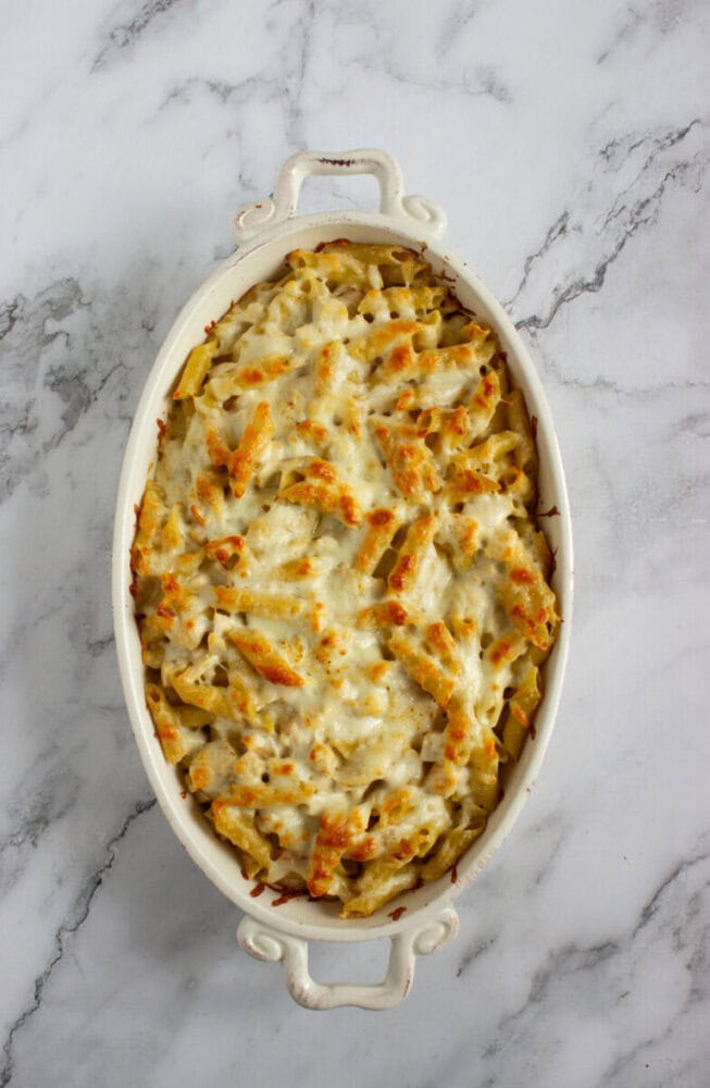 A white oval baking dish filled with baked pasta topped with melted, slightly browned cheese, set on a marble surface.
