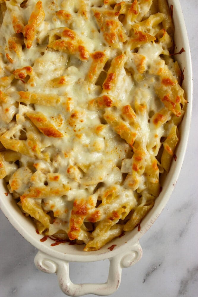 A baked pasta casserole with penne noodles covered in melted and lightly browned cheese in a white ceramic dish on a marble surface.