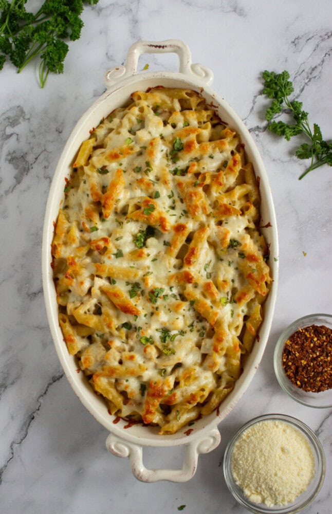 Baked pasta casserole with melted cheese on top in an oval dish, garnished with chopped herbs. Bowls of grated cheese and red pepper flakes are nearby on a marble surface.