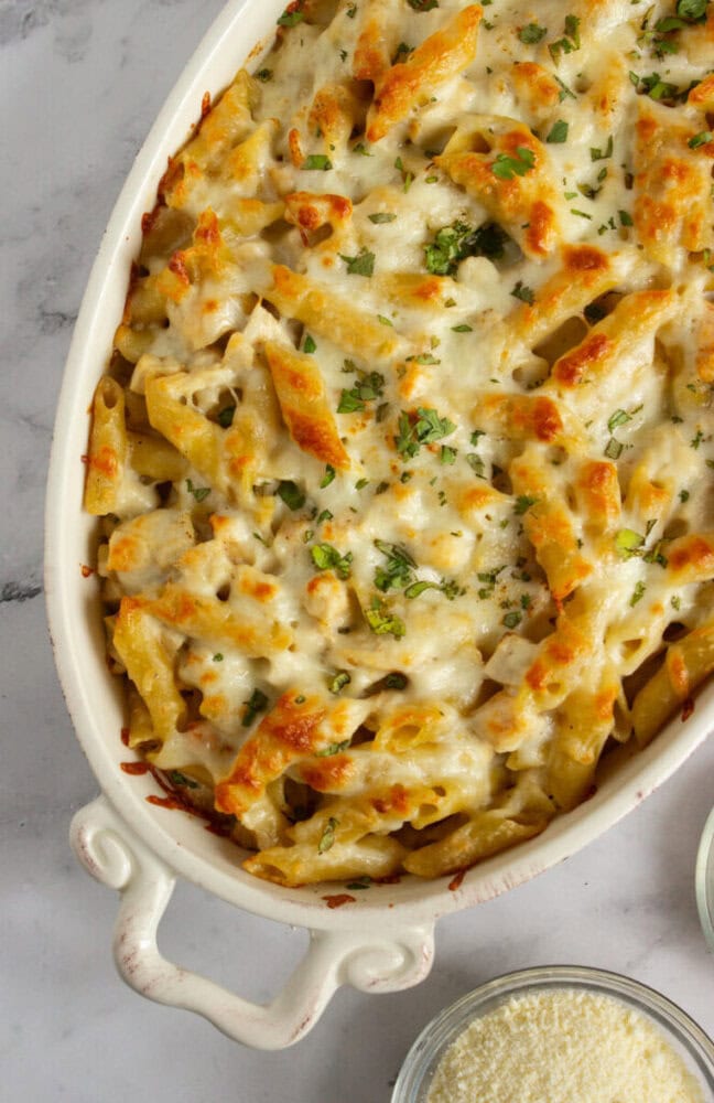 Baked penne pasta casserole topped with melted cheese and chopped herbs in a white oval dish, placed on a marble surface.