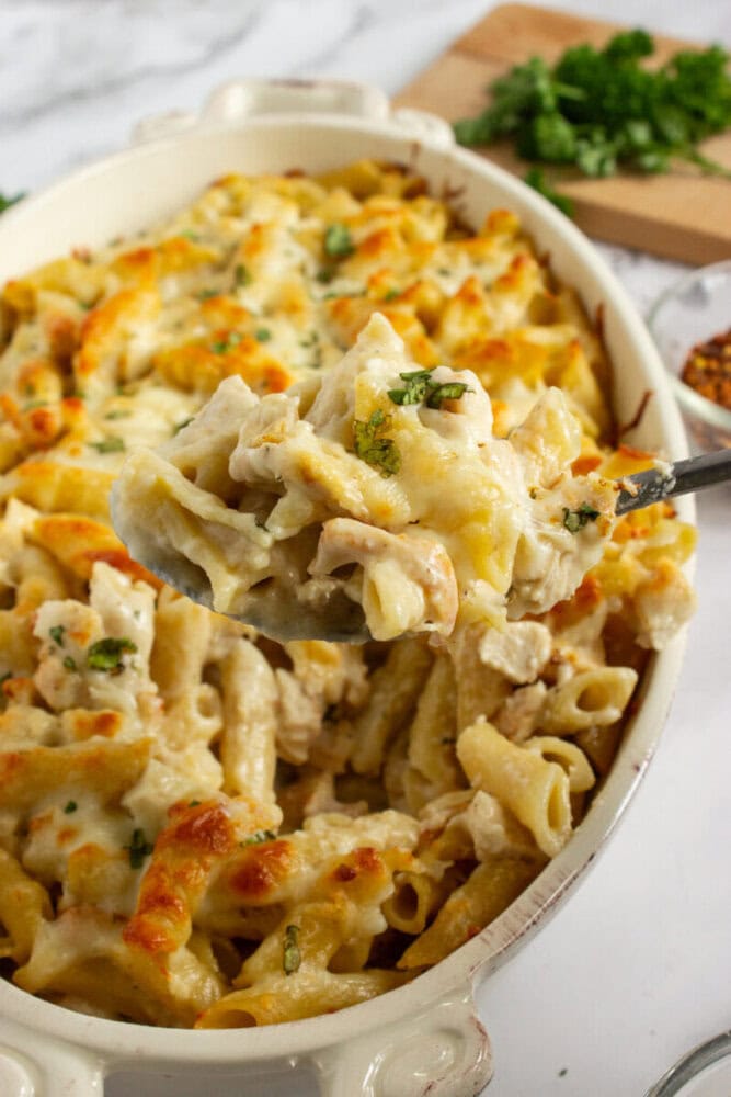 A serving spoon holds a portion of baked pasta with melted cheese and herbs above a casserole dish filled with the same pasta.