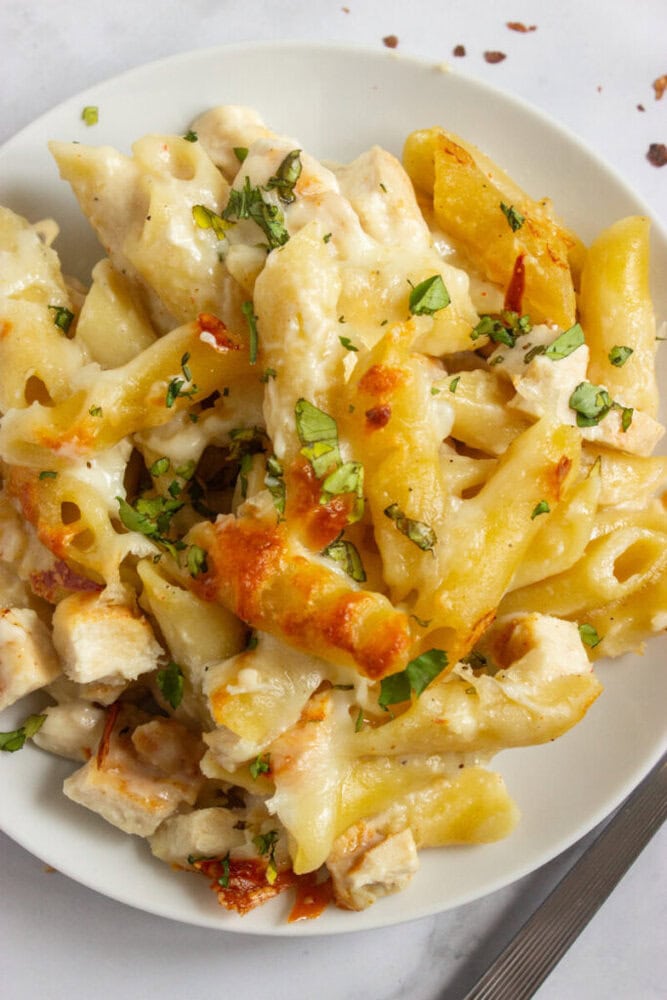 A plate of baked penne pasta with pieces of chicken, melted cheese, and fresh herbs sprinkled on top.