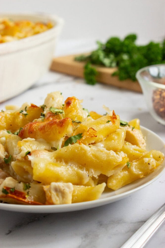 A plate of baked ziti pasta with melted cheese and herbs, served on a white plate, with fresh parsley and red pepper flakes in the background.