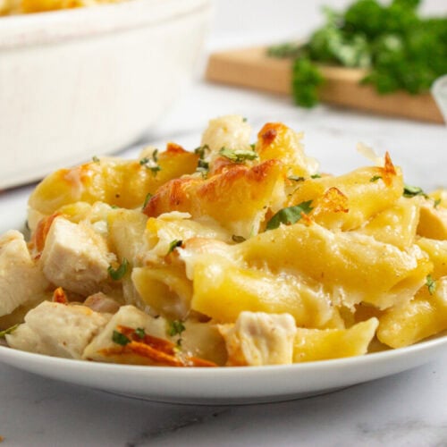 A plate of baked penne pasta with pieces of chicken, melted cheese, and chopped herbs, on a white surface with parsley and a spice bowl in the background.