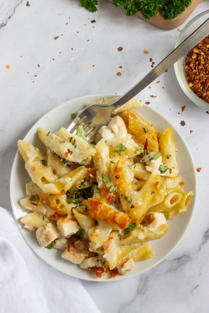 A plate of baked penne pasta with chunks of chicken, creamy sauce, chopped herbs, and a fork, garnished with red pepper flakes.