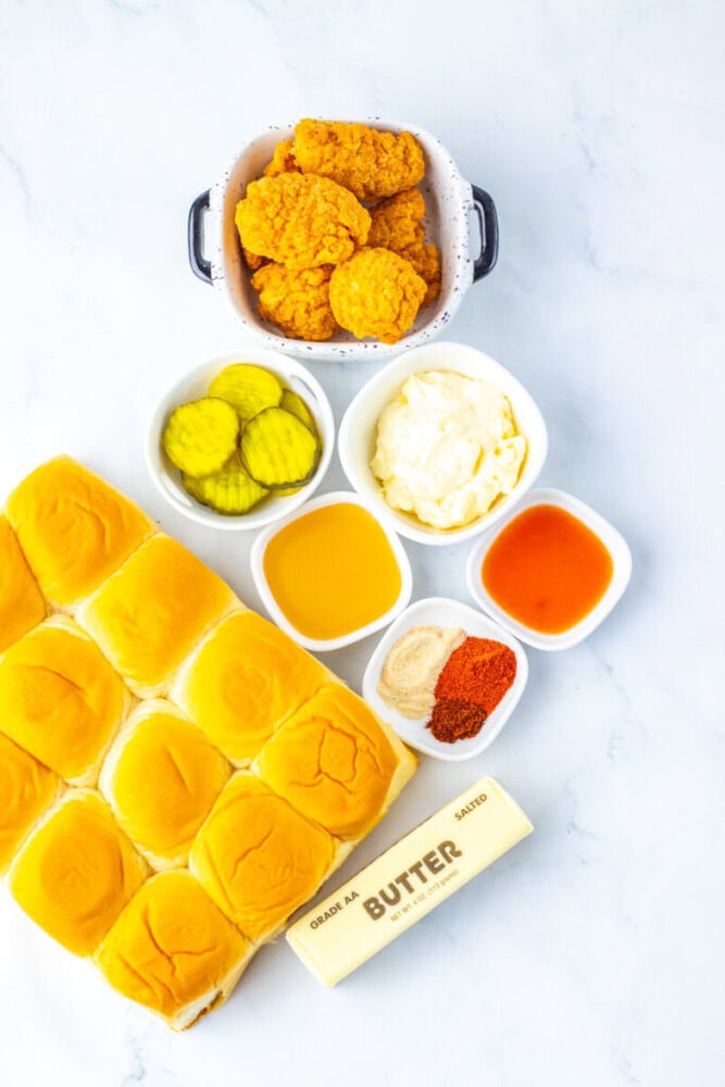 A spread of slider buns, breaded chicken patties, pickle slices, mayonnaise, melted butter, hot sauce, spice mixture, and a stick of butter on a white surface.