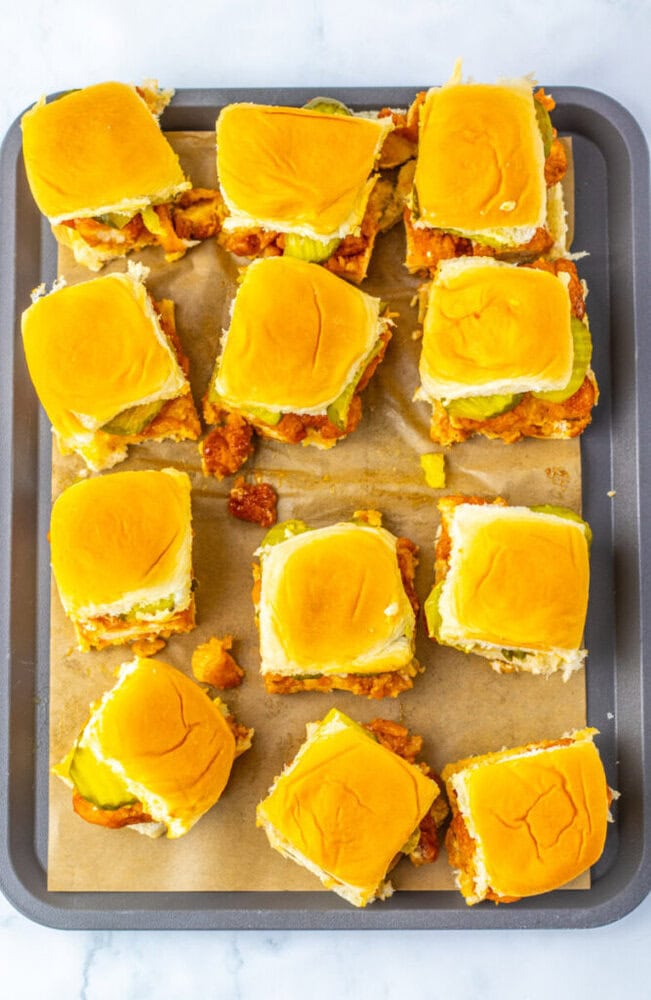 A baking sheet lined with parchment paper holds twelve chicken slider sandwiches topped with pickles and golden buns, arranged in neat rows.