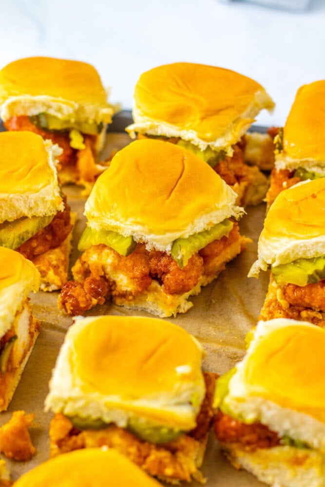 Close-up of several crispy chicken sliders with pickles on soft dinner rolls, arranged on a parchment-lined baking sheet.