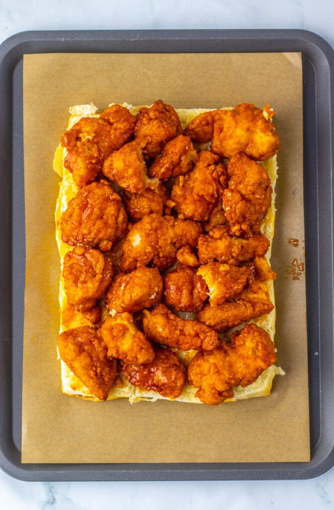 A baking tray with parchment paper holds a rectangular piece of bread topped with saucy, breaded fried chicken pieces.