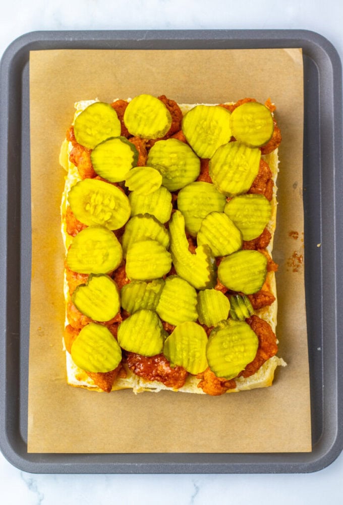 Rectangular sandwich on parchment paper topped with sauced chicken pieces and layers of crinkle-cut pickles, on a baking tray.