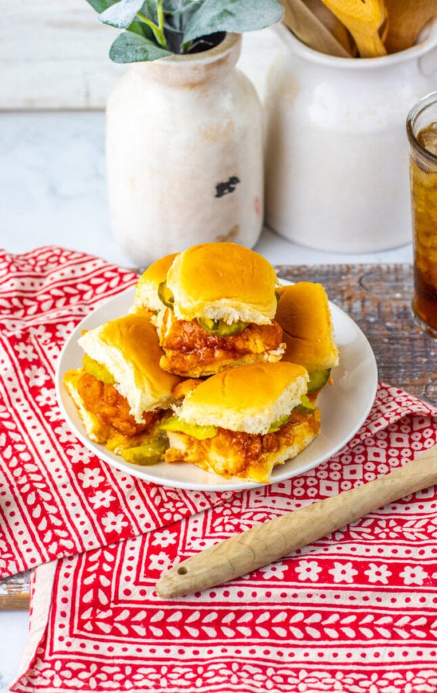 A plate with four chicken sliders on white buns, set on a red and white patterned cloth, with jars and a glass of iced drink in the background.