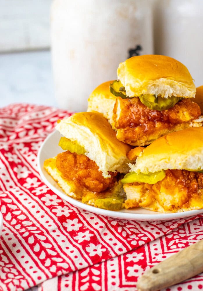 A plate of fried chicken sliders with pickles on soft buns, placed on a red and white patterned cloth.