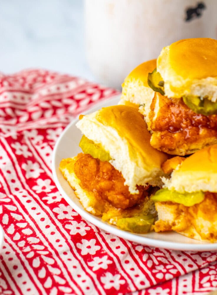 A plate of fried chicken sliders with pickles on soft buns, placed on a red and white patterned cloth.